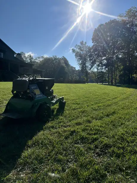 A stand-on aerifier with a fertilizer hopper crossing a Northwest Georgia lawn into the sun