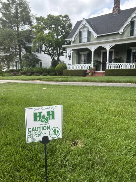 An H&H caution sign on a just-treated lawn in front of a wraparound porch